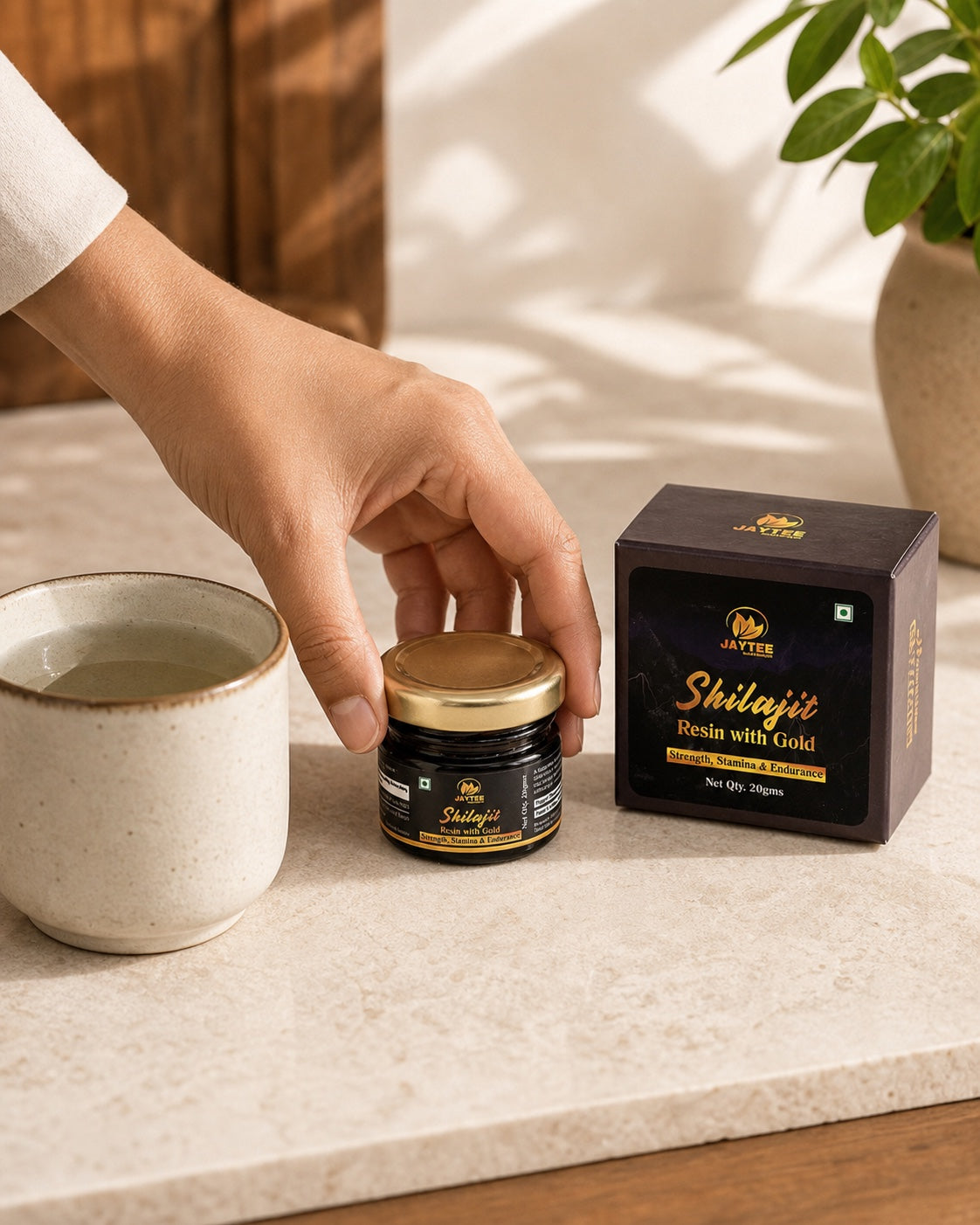 Adult hand positioning a closed Shilajit Resin with Gold jar beside a ceramic cup of lukewarm water